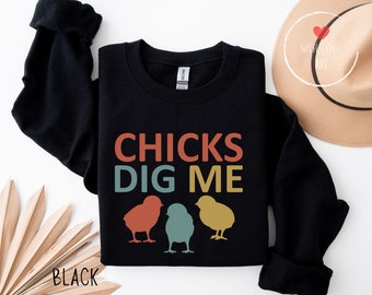 Może przedstawiać: Czarna bluza z napisem "CHICKS DIG ME" w kolorze pomarańczowym, turkusowym i żółtym. Poniżej znajdują się trzy sylwetki piskląt w tych samych kolorach. Na dole nadrukowano słowo "BLACK".