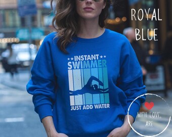 Op de afbeelding: Koningsblauw sweatshirt met de tekst "INSTANT SWIMMER JUST ADD WATER" en een zwemmerafbeelding. Het sweatshirt heeft een effen koningsblauwe kleur.