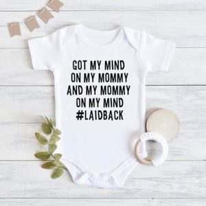 May include: White baby onesie with black text that reads "Got my mind on my mommy and my mommy on my mind #laidback"