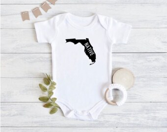 Könnte beinhalten: Weißer Baby-Strampler mit einer schwarzen Umrandung des Bundesstaates Florida und dem Wort "NATIVE" innerhalb des Bundesstaates.