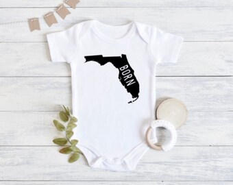 Könnte beinhalten: Weißer Baby-Strampler mit einer schwarzen Umrandung des Bundesstaates Florida und dem Wort "BORN" vertikal innerhalb des Bundesstaates geschrieben.