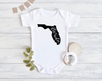 Könnte beinhalten: Weißer Baby-Strampler mit einer schwarzen Umrandung des Bundesstaates Florida und dem Wort "Florida" in Kursivschrift.