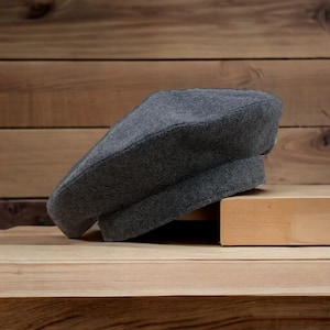 May include: A grey beret hat made of felt, sitting on a wooden surface.