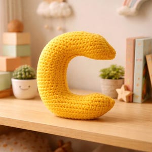 May include: A vibrant yellow, crocheted letter 'C' rests on a wooden shelf. The handmade item is the main focus, with a textured surface and curved form. The background features decorative elements such as a small potted plant and books.