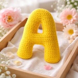 May include: A bright yellow crocheted letter "A" sits in a wooden tray. The handmade letter is made of yarn and has a textured appearance. Pink and white flowers are in the background.