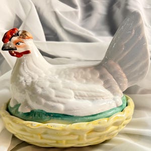 May include: A white ceramic hen-shaped container with a red comb and beak, sitting in a yellow basket-like base with green accents. The container is likely used for storing eggs or as a decorative piece.