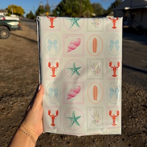 May include: A package wrapped in white paper with a repeating pattern of stamps featuring a red lobster, a blue bow, a pink seashell, a green starfish, a surfboard, and flowers. The package is held in a person's hand.