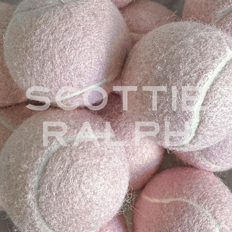 May include: A collection of fuzzy, light pink tennis balls with white seams. The text SCOTTIE RALPH is overlaid in white capital letters across the centre of the cluster of balls, which are arranged in a dense, overlapping pile.