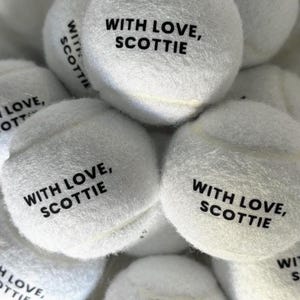 May include: A collection of white fuzzy tennis balls, each featuring the black printed text "WITH LOVE, SCOTTIE" on the surface. The balls are arranged in a cluster, showcasing the soft texture and the consistent branding across the items.