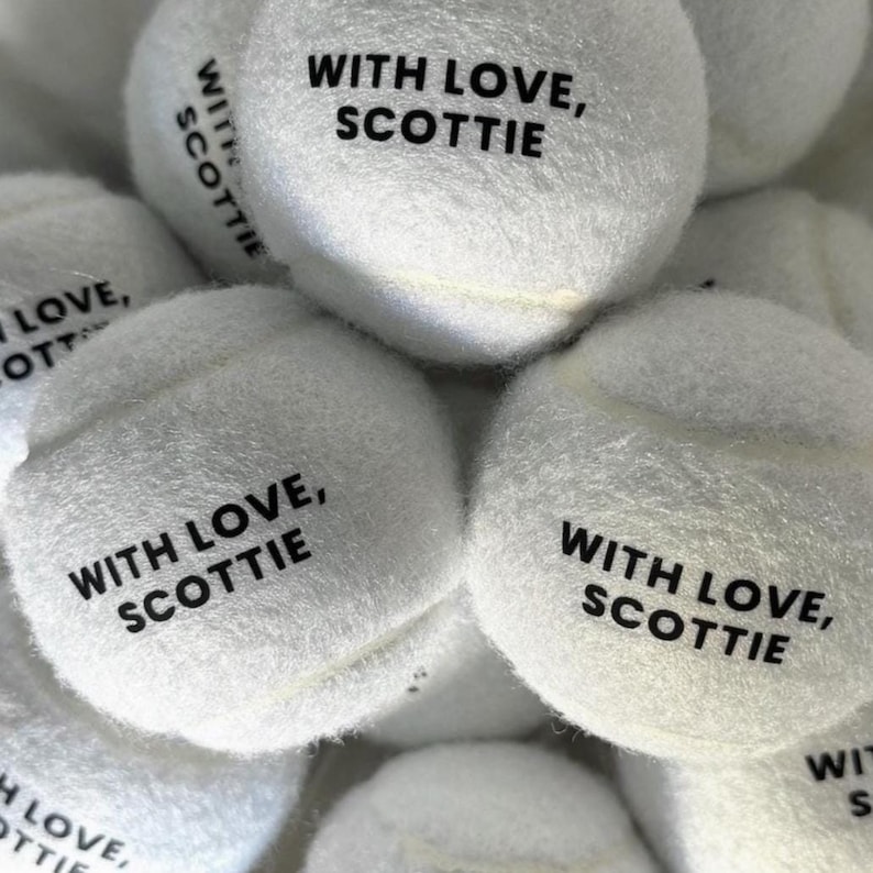 May include: A collection of white fuzzy tennis balls, each featuring the black printed text "WITH LOVE, SCOTTIE" on the surface. The balls are arranged in a cluster, showcasing the soft texture and the consistent branding across the items.