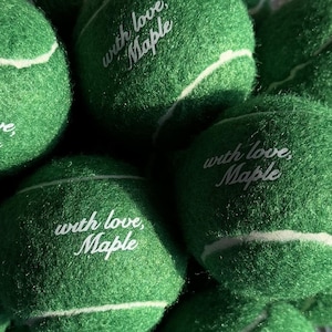May include: A collection of bright green tennis balls with a fuzzy texture and white curved seams. Each ball features the white printed text "with love, Maple" in a cursive script font, centred on the surface of the ball.