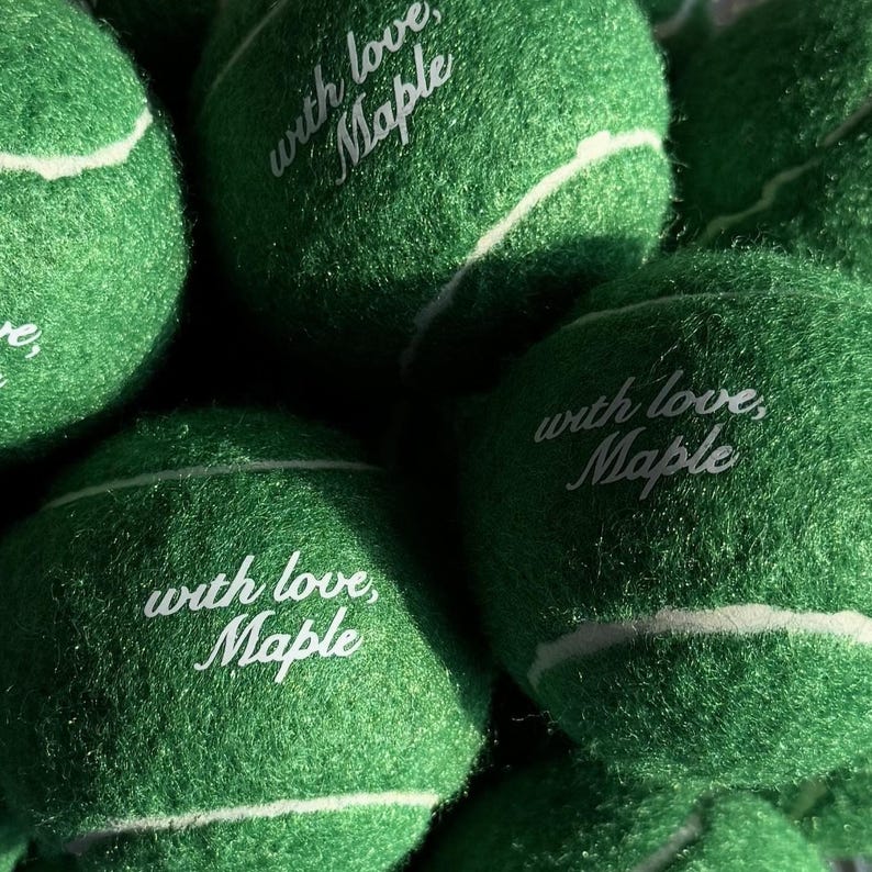 May include: A collection of bright green tennis balls with a fuzzy texture and white curved seams. Each ball features the white printed text "with love, Maple" in a cursive script font, centred on the surface of the ball.