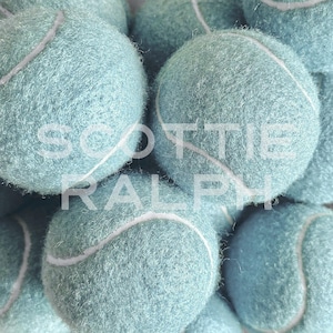 May include: A collection of fuzzy, light blue tennis balls with white seams. The text SCOTTIE RALPH is overlaid in white capital letters across the centre of the image, partially obscuring the textured surface of the spherical objects.