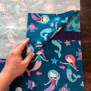 May include: A teal blue fabric with a mermaid pattern. The fabric has a purple trim and is folded over a light blue fabric with a mermaid pattern.