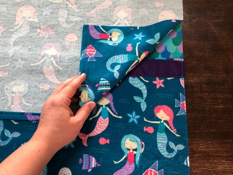 May include: A teal blue fabric with a mermaid pattern. The fabric has a purple trim and is folded over a light blue fabric with a mermaid pattern.