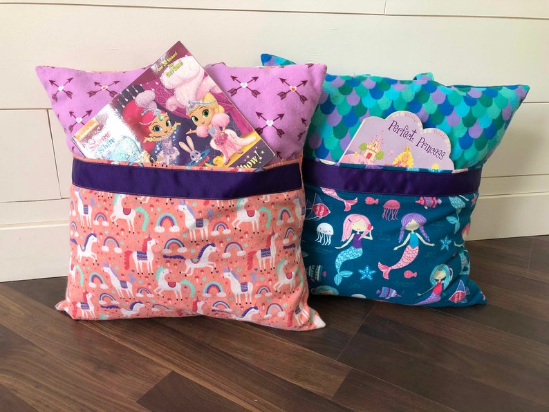 May include: Two colorful pillow book holders with pockets for books. The pillow on the left has a pink and white unicorn pattern and a purple ribbon. The pillow on the right has a teal and blue mermaid pattern and a purple ribbon. Both pillows have books tucked into the pockets.