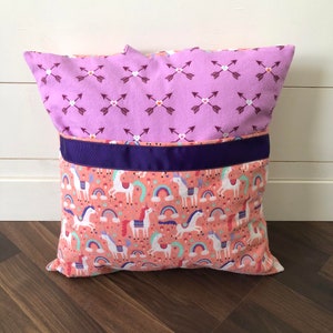 May include: A purple and pink pillow with a pattern of arrows and hearts on one side and a pattern of unicorns and rainbows on the other side. The pillow has a purple ribbon around the middle.