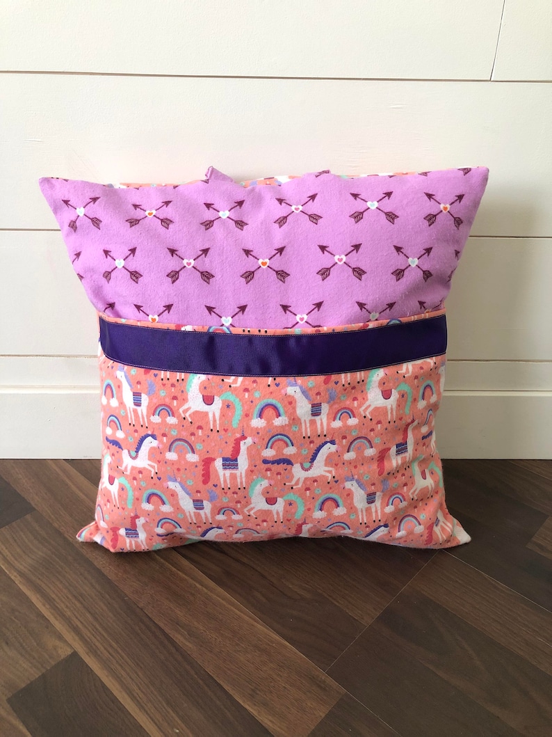 May include: A purple and pink pillow with a pattern of arrows and hearts on one side and a pattern of unicorns and rainbows on the other side. The pillow has a purple ribbon around the middle.