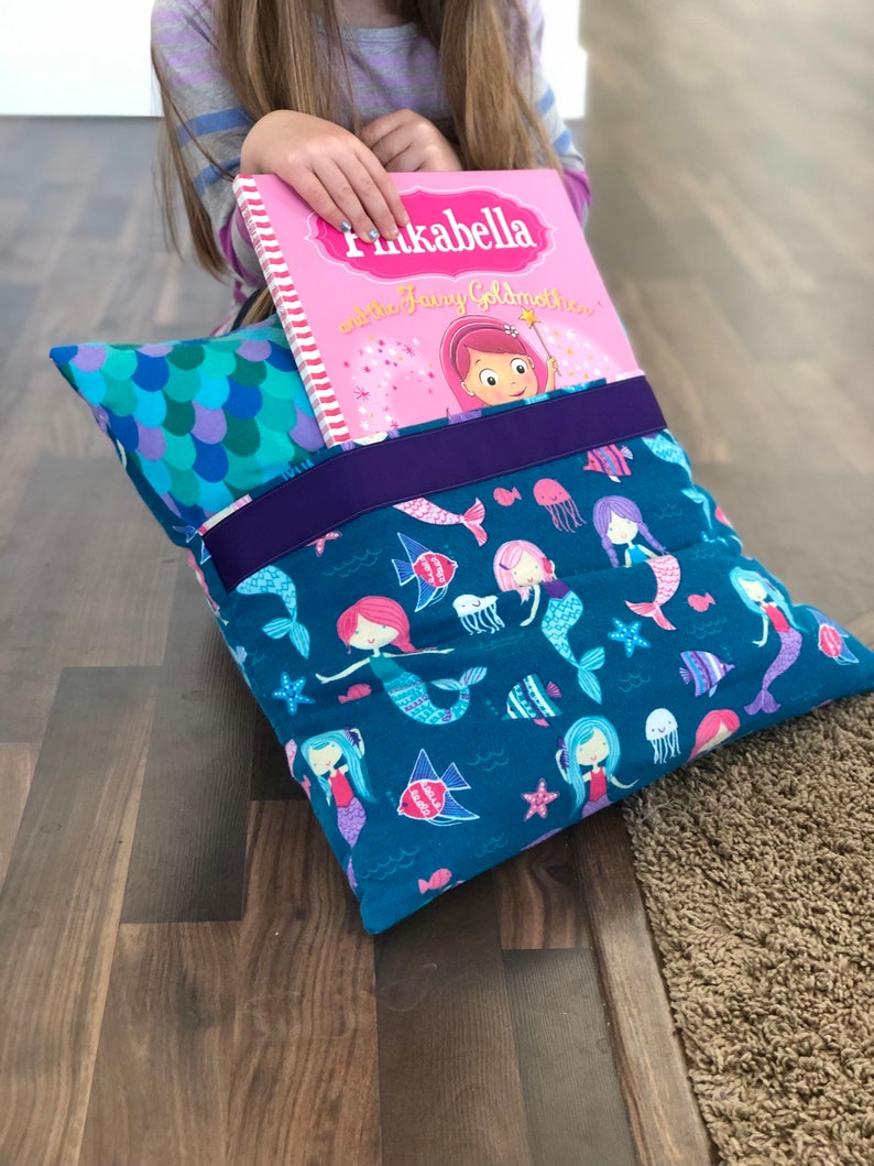 May include: A teal blue pillow with a mermaid pattern and a purple trim. The pillow has two pockets, one on each side, and a book titled "Pinkabella and the Fairy Godmother" is tucked into the top pocket.