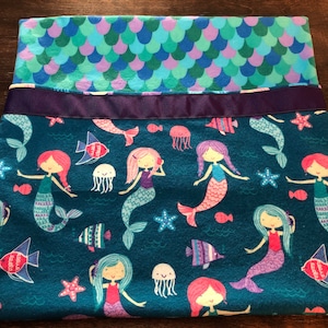 May include: A teal blue fleece blanket with a mermaid pattern. The blanket features mermaids with pink, purple, and blue tails, starfish, jellyfish, and fish. The blanket has a purple ribbon sewn across the top.