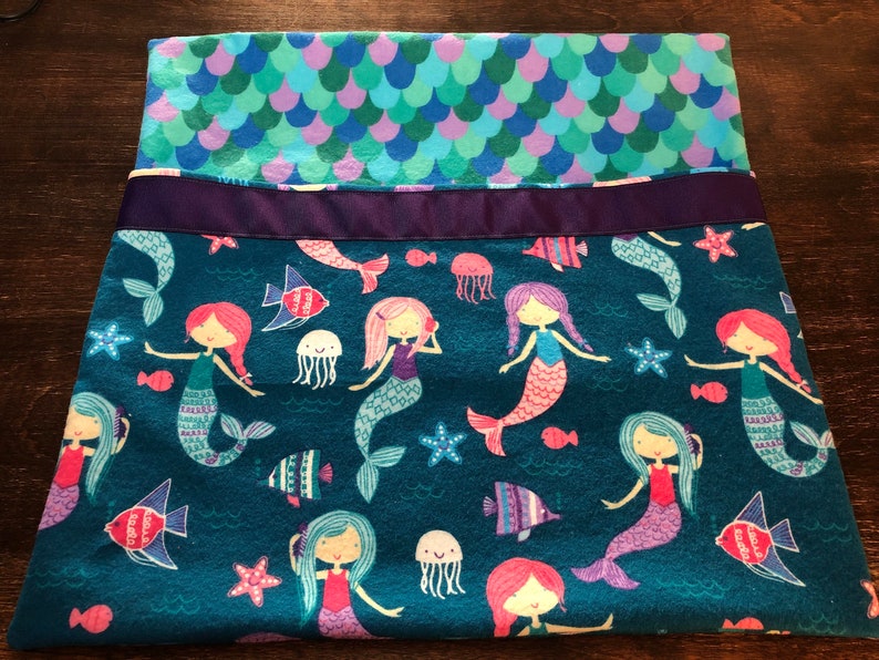 May include: A teal blue fleece blanket with a mermaid pattern. The blanket features mermaids with pink, purple, and blue tails, starfish, jellyfish, and fish. The blanket has a purple ribbon sewn across the top.