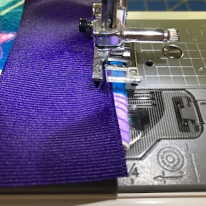 May include: A close-up of a sewing machine needle stitching together a purple ribbon and a blue and pink fabric with a starfish design.