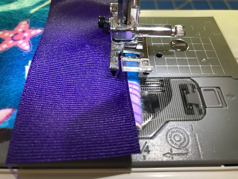 May include: A close-up of a sewing machine needle stitching together a purple ribbon and a blue and pink fabric with a starfish design.