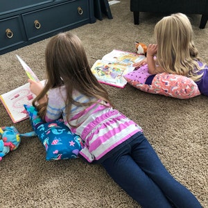May include: Two children are lying on the floor reading books. The girl on the left is using a blue and green pillow with a mermaid design. The girl on the right is using a pink pillow with a unicorn design. Both girls are wearing casual clothing.