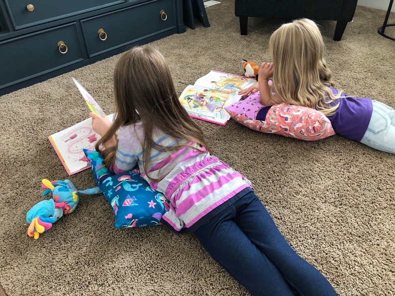 May include: Two children are lying on the floor reading books. The girl on the left is using a blue and green pillow with a mermaid design. The girl on the right is using a pink pillow with a unicorn design. Both girls are wearing casual clothing.
