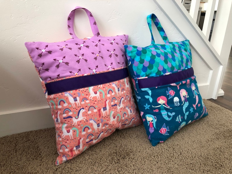 May include: Two colorful pillow-like bags with handles. One has a pink and purple fabric with a unicorn and rainbow pattern. The other has a blue and green fabric with a mermaid and fish pattern.