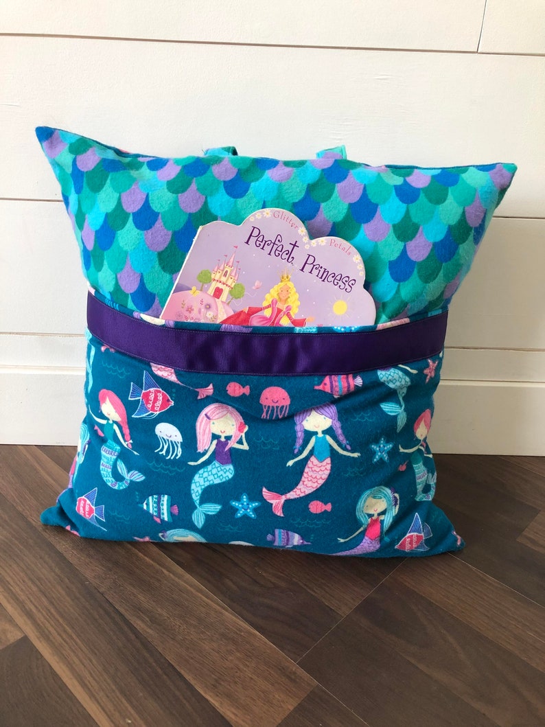 May include: A teal blue pillow with a pocket on the front. The pocket has a purple ribbon and a book titled "Glitter Petals Perfect Princess" is tucked inside. The pillow has a mermaid pattern on the front and a scale pattern on the back.