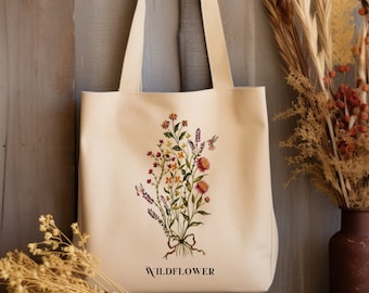 May include: Cream-colored tote bag featuring a colorful embroidered wildflower bouquet with a hummingbird. The word "WILDFLOWER" is printed in black at the bottom. The bag has two long handles.