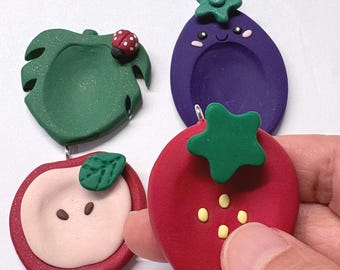 May include: A collection of four fruit and vegetable shaped charms made of polymer clay. The set includes a green leaf with a ladybug, a purple eggplant with a face, a red apple slice, and a red strawberry with a green star-shaped top.