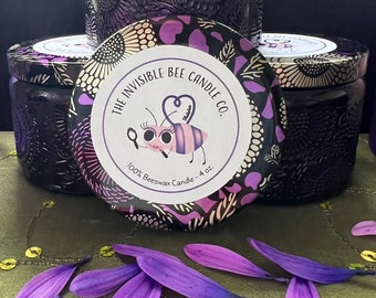 May include: Three purple and gold patterned glass candle jars with lids. The middle jar has a label that reads "The Invisible Bee Candle Co." with a cartoon bee illustration and the text "100% Beeswax Candle - 4 oz."