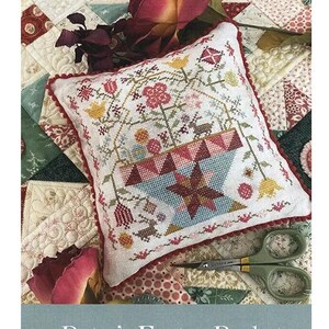 May include: A cross-stitch pattern for "Betsy's Easter Basket" featuring a floral basket design with a star. The pattern is on a white pillow with a red border, and the text "Pansy Patch Quilts and Stitchery" is visible. Scissors are in the foreground.