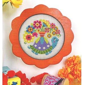 May include: A cross-stitch pattern featuring a turtle carrying a vase of colorful flowers, set in a round, orange, scalloped frame. The text "flower friends myrtle" is at the top, and "satsuma street" is at the bottom.