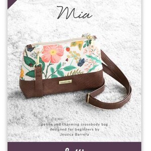 May include: A floral print crossbody bag with a brown leather strap and a zipper closure. The bag is designed by Jessica Barrera and is called "Mia".