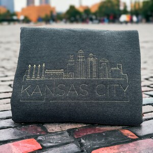 May include: A dark gray sweatshirt with a gold embroidered outline of the Kansas City skyline and the text "KANSAS CITY".