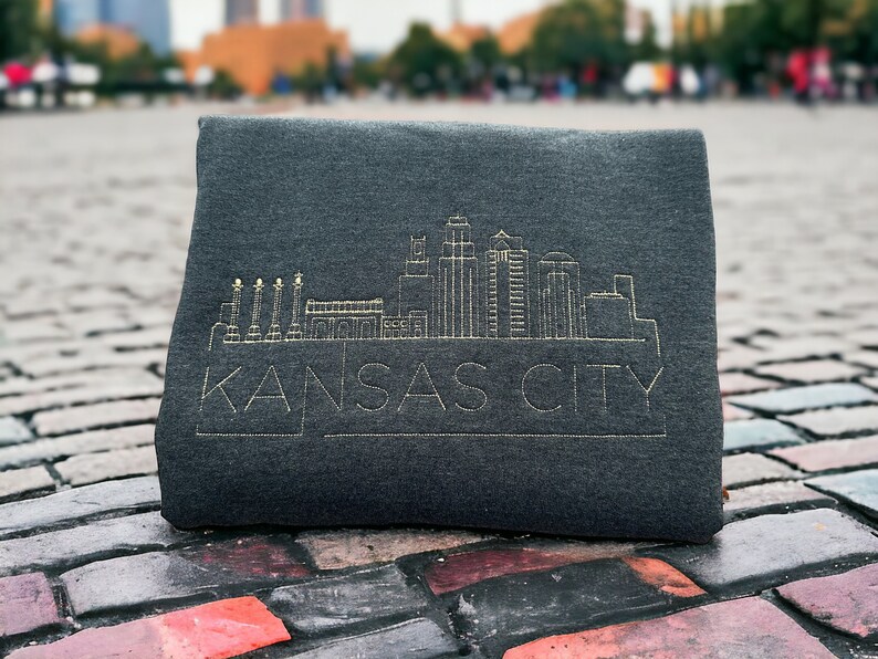 May include: A dark gray sweatshirt with a gold embroidered outline of the Kansas City skyline and the text "KANSAS CITY".