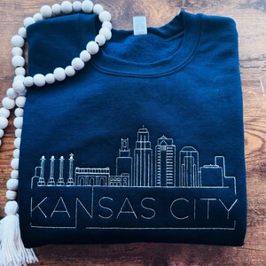 May include: A navy blue sweatshirt with a white embroidered skyline of Kansas City. The sweatshirt has a crew neck and long sleeves.