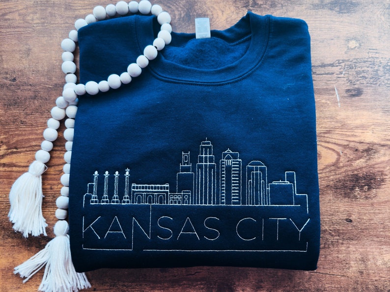 May include: A navy blue sweatshirt with a white embroidered skyline of Kansas City. The sweatshirt has a crew neck and long sleeves.