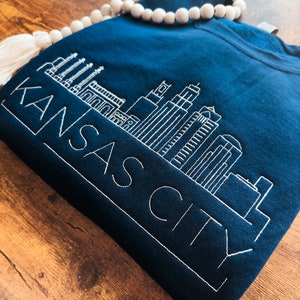 May include: A navy blue sweatshirt with a white embroidered design of the Kansas City skyline and the text "KANSAS CITY".