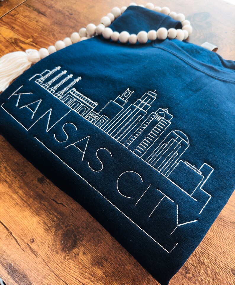 May include: A navy blue sweatshirt with a white embroidered design of the Kansas City skyline and the text "KANSAS CITY".