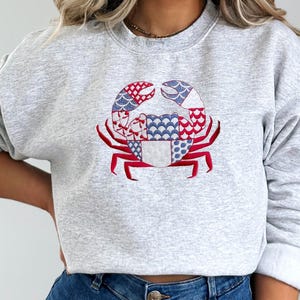 Może przedstawiać: Jasnoszara bluza z haftowanym wzorem kraba. Krab jest ozdobiony czerwonymi, białymi i niebieskimi wzorami. Bluza ma długie rękawy i okrągły dekolt. Wzór jest umieszczony na środku przodu.