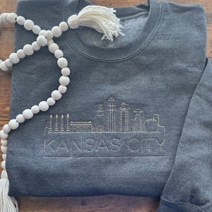 May include: A dark gray sweatshirt with a gold embroidered design of the Kansas City skyline. The text "KANSAS CITY" is below the skyline.