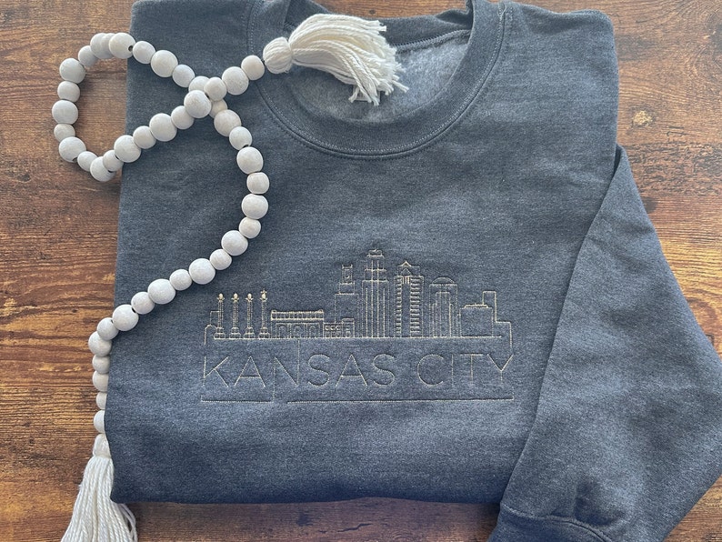 May include: A dark gray sweatshirt with a gold embroidered design of the Kansas City skyline. The text "KANSAS CITY" is below the skyline.
