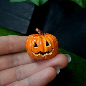 Può includere: Una miniatura di zucca jack-o'-lantern arancione con un occhio triangolare intagliato e una bocca frastagliata. La zucca presenta un gambo marrone strutturato sulla parte superiore e una finitura lucida, con dettagliate creste verticali.