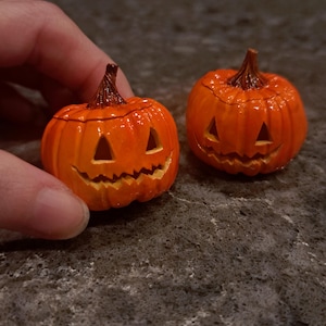May include: Two small, glossy orange jack-o'-lantern figurines with carved triangular eyes and jagged mouths. Each pumpkin features a textured brown stem at the top and a rounded, ribbed body. The pumpkins are crafted with a smooth, shiny finish.