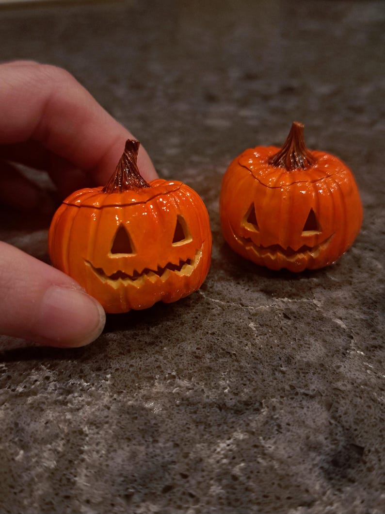 May include: Two small, glossy orange jack-o'-lantern figurines with carved triangular eyes and jagged mouths. Each pumpkin features a textured brown stem at the top and a rounded, ribbed body. The pumpkins are crafted with a smooth, shiny finish.