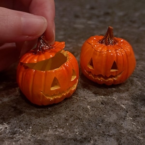 May include: Two small orange jack-o'-lanterns with carved triangular eyes and jagged mouths. One pumpkin is shown with its lid slightly lifted, revealing a hollow interior, while the other pumpkin remains closed with a dark brown stem on top.